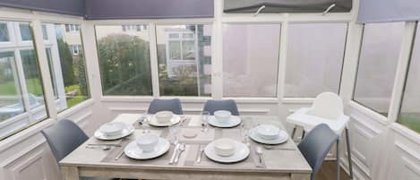 Un espace repas aménagé dans une véranda, avec une table dressée pour quatre personnes, des chaises et de grandes fenêtres.