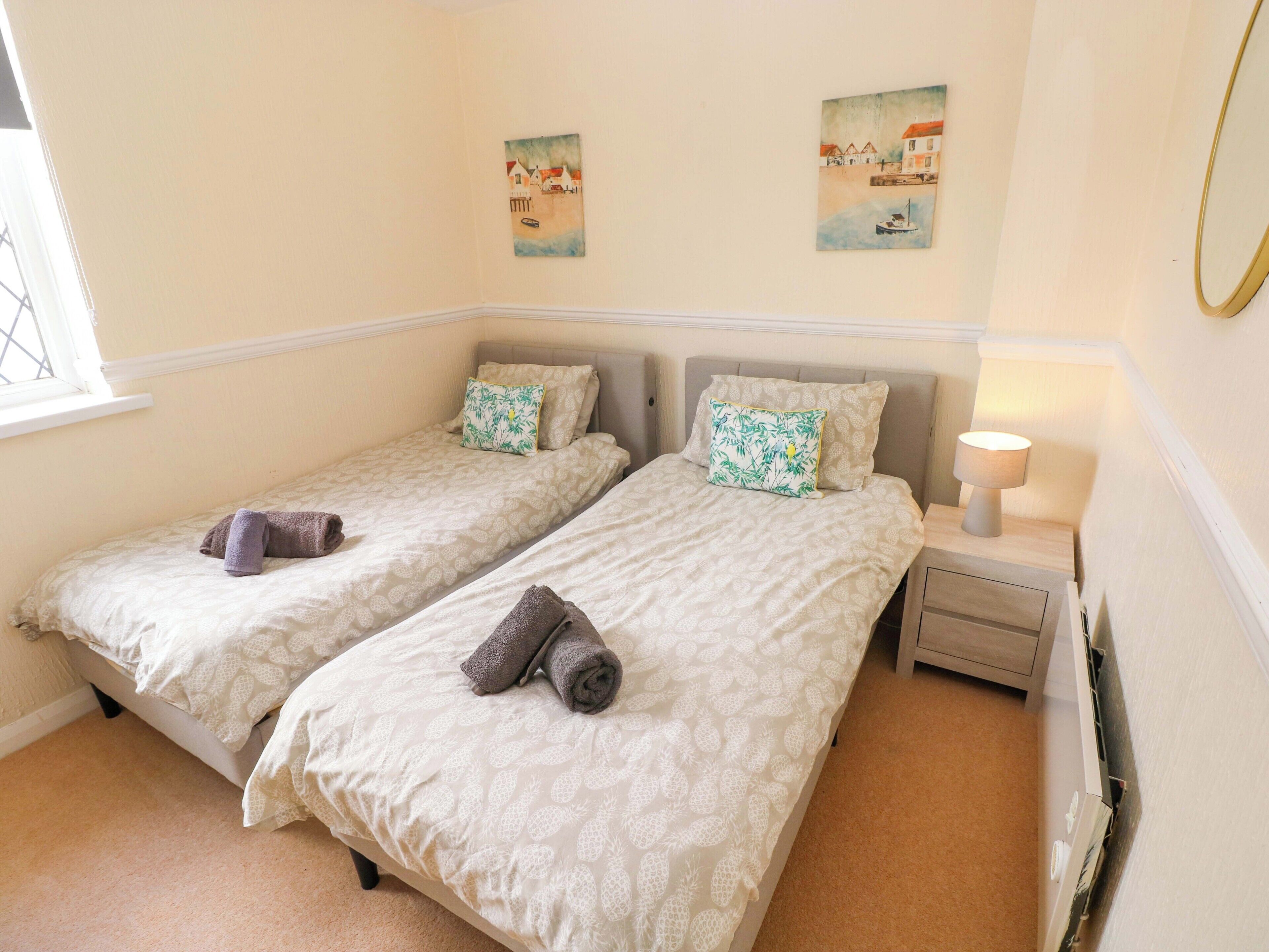 A double room with two beds, each with a pillow and a rolled towel. There is a nightstand with a lamp, a window, and two wall art pieces.