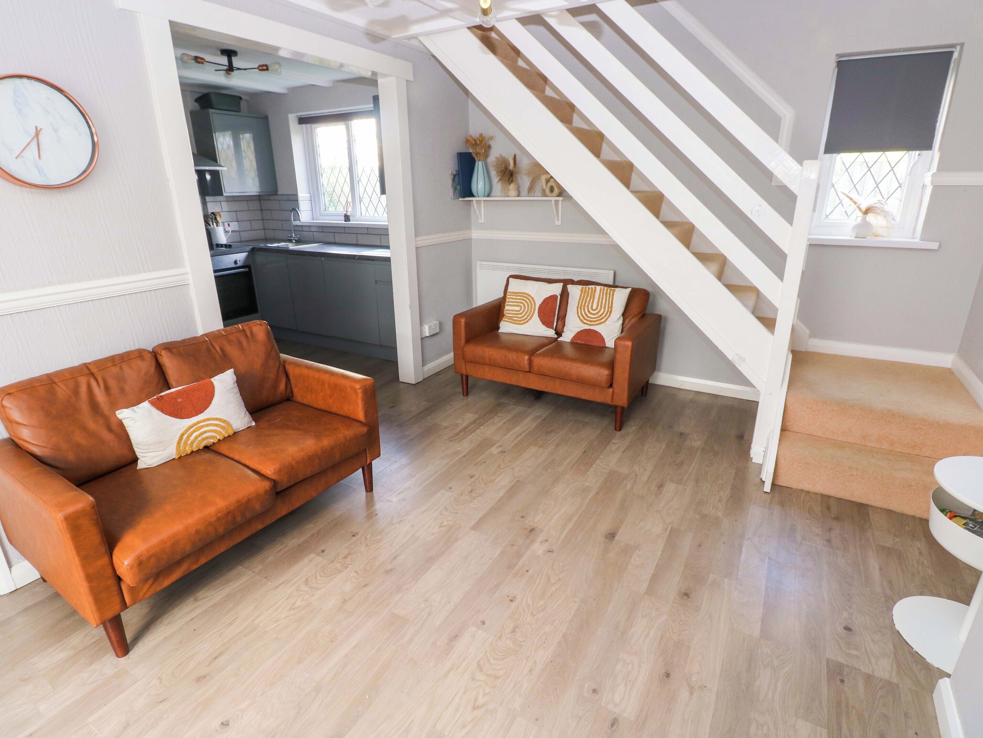 A living room with a brown leather sofa and a smaller sofa with patterned cushions, a staircase, and a kitchen in the background.