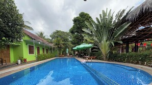 Une piscine dont le bord est revêtu de carreaux, entourée de verdure et bordée d’une structure à toit de chaume.