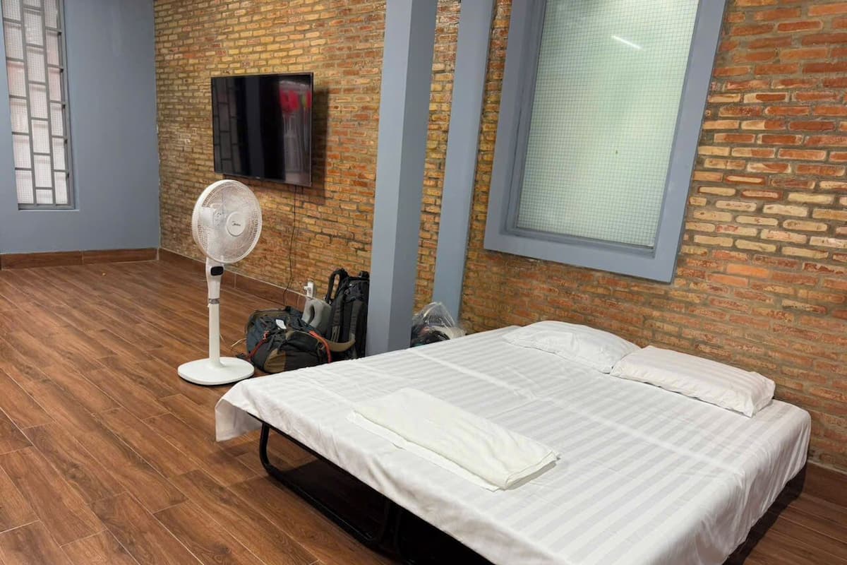 Een hotelkamer met een bakstenen muur, een bed met wit beddengoed, een staande ventilator, een aan de muur bevestigde televisie en een raam met een rasterpatroon.