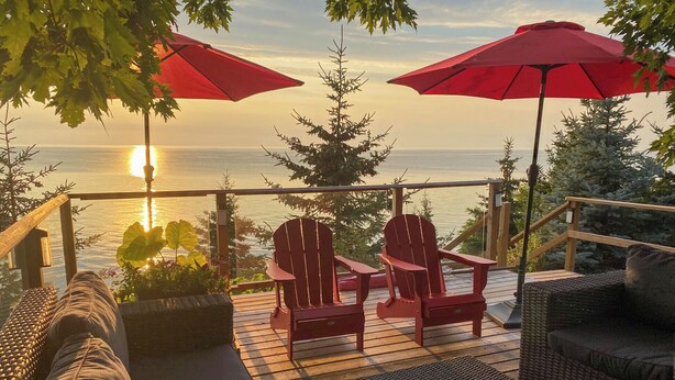 Terraza con mobiliario de mimbre, dos sillas Adirondack y sombrillas rojas con vista a una masa de agua.