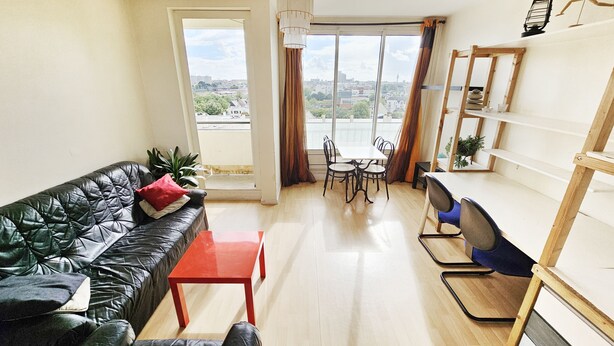 A living room with a leather sofa, a red table, a dining area with chairs, and a view through a large window. - Pleasant, bright T3 on top floor (8), not overlooked, close to parks/services (Rennes)