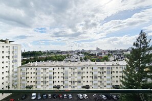 Ein Hochhaus mit mehreren Balkonen, ein Parkplatz mit verschiedenen Autos und ein Blick auf eine Stadtlandschaft mit Gebäuden und Bäumen unter einem teils bewölkten Himmel.