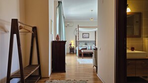 A hallway leading to a bedroom with a four-poster bed, a wooden chair, a small table with a lamp, and a mirror in the bathroom.