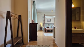 A hallway leading to a bedroom with a four-poster bed, a wooden chair, a small table with a lamp, and a mirror in the bathroom.