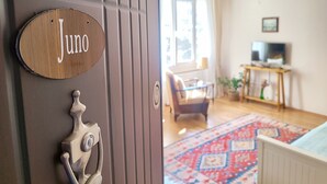 A wooden door with a nameplate reading 'Juno' and a keyhole. Inside, there is a living room with a sofa, a TV, a wooden table, and a patterned rug.