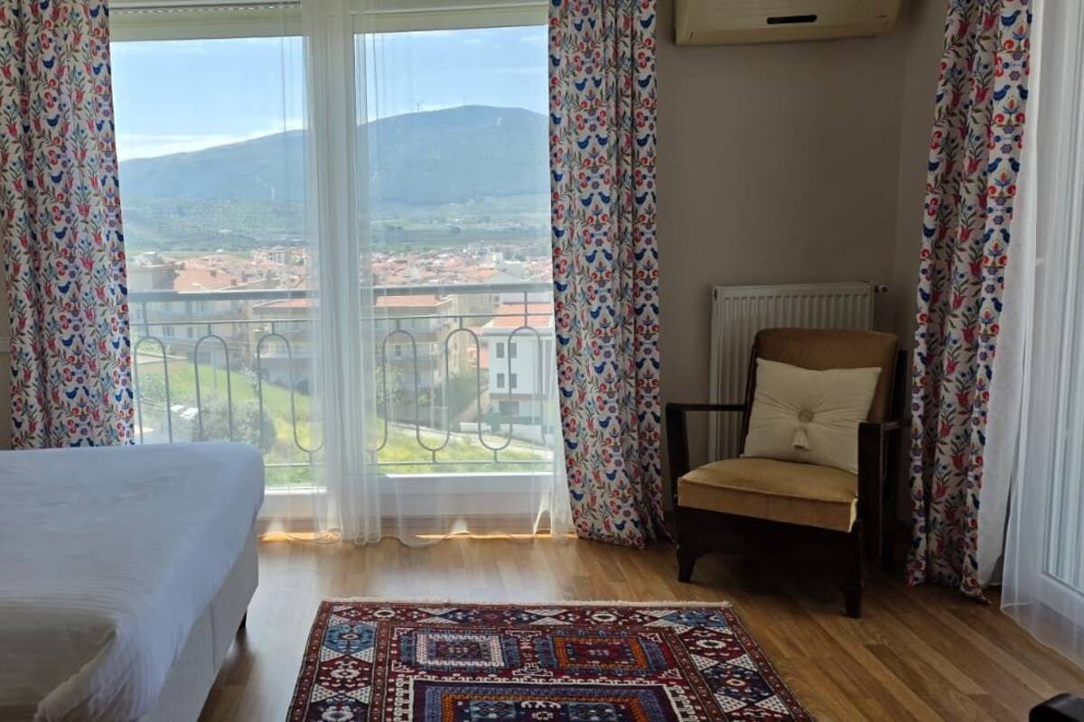 Une chambre avec un grand lit, une chaise, une vue sur la ville et les montagnes, et un tapis à motifs.