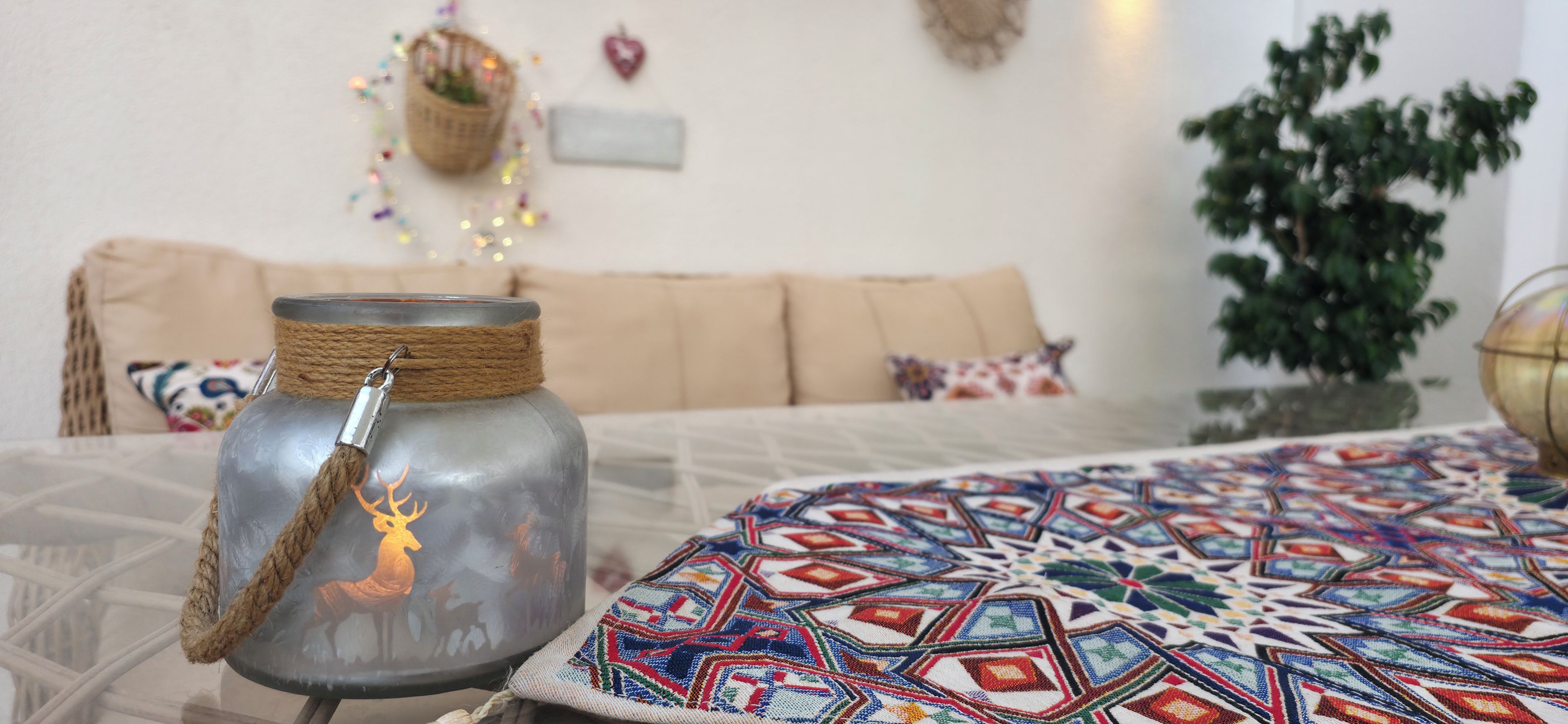Quarto com pote de vidro e alça de corda, silhueta de veado, toalha de mesa estampada, sofá com almofadas, luminária na parede e planta em vaso.