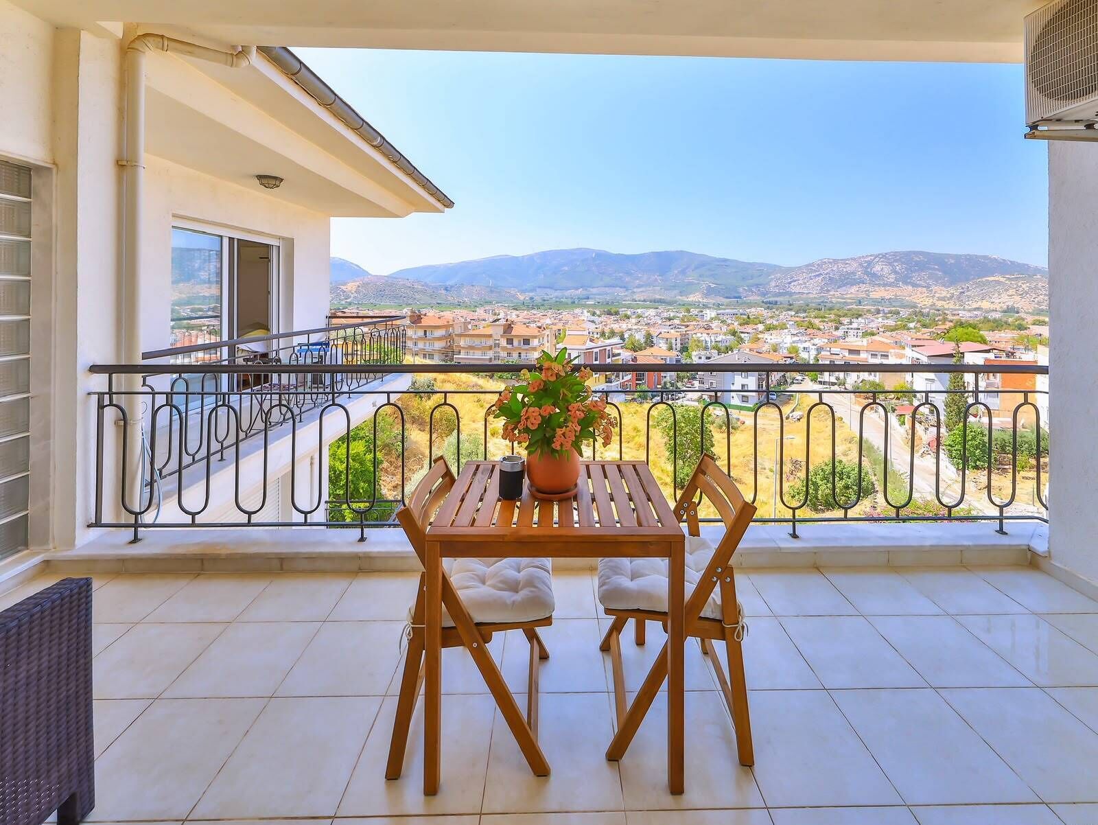 A balcony with a table and chairs, a potted plant, and a view of a town and mountains.
