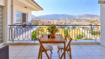 A balcony with a table and chairs, a potted plant, and a view of a town and mountains.