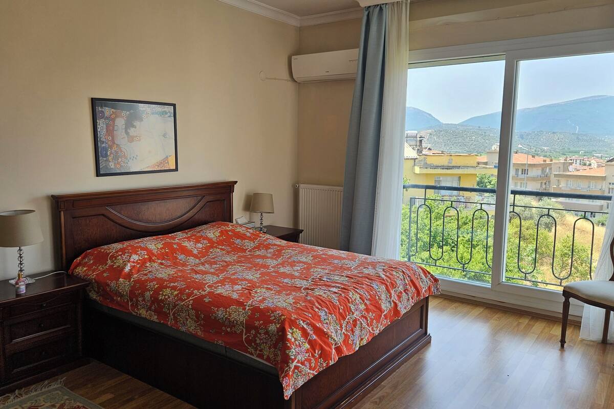 Une chambre à coucher comprenant un lit, une chaise, une table de chevet, une lampe, un tableau et une fenêtre donnant sur des bâtiments et des montagnes.