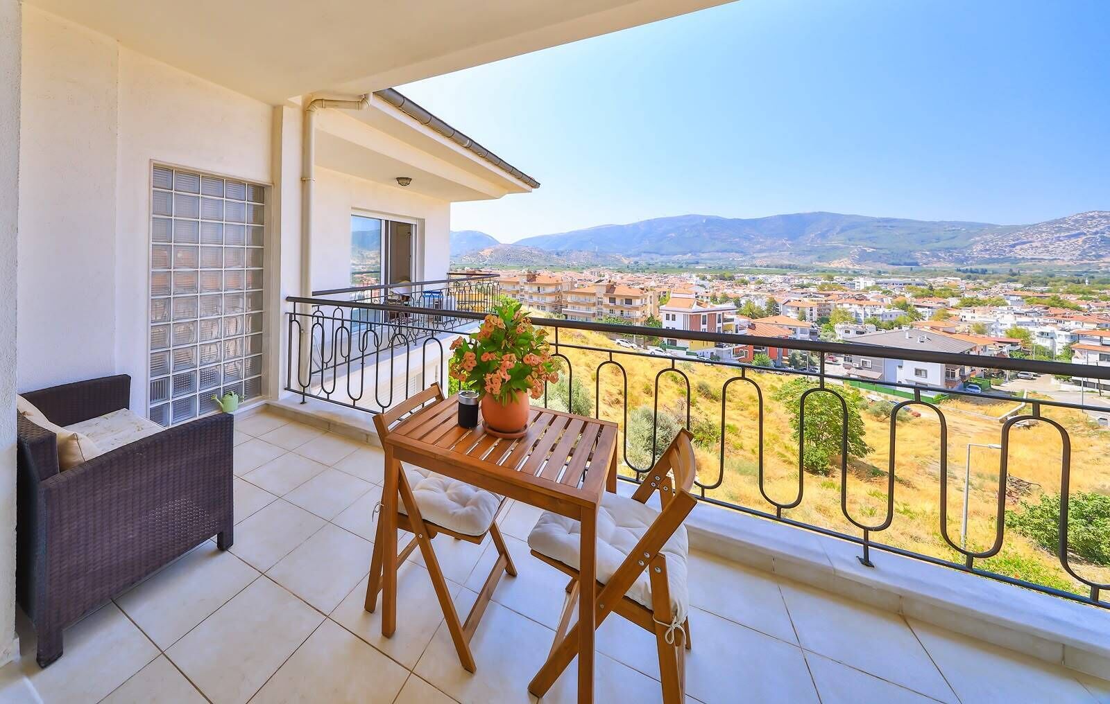 A balcony with a table and chairs, a potted plant, and a view of a town and mountains.