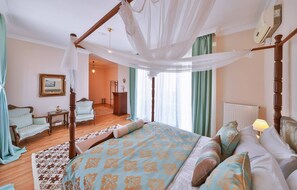 A bedroom with a four-poster bed, a sitting area with a chair and a table, and a view of the hallway. - Ephesus Suites (Selçuk)