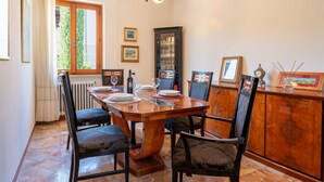 Un comedor con una mesa de madera, sillas, una araña, un ventanal con cortinas y un aparador con cristalería.