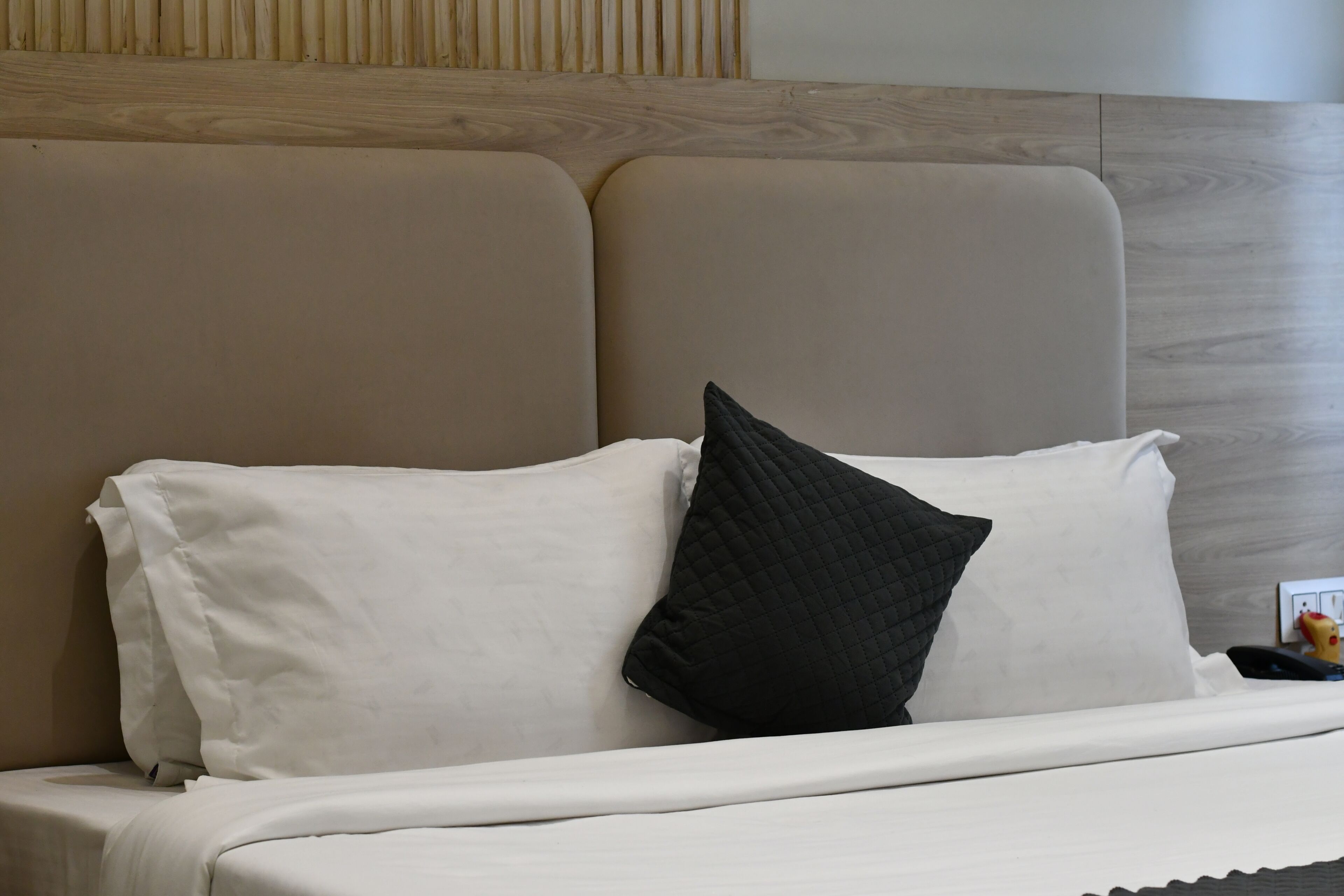 A bed with white bedding and a black pillow, a wooden headboard, and a small yellow object on the nightstand.