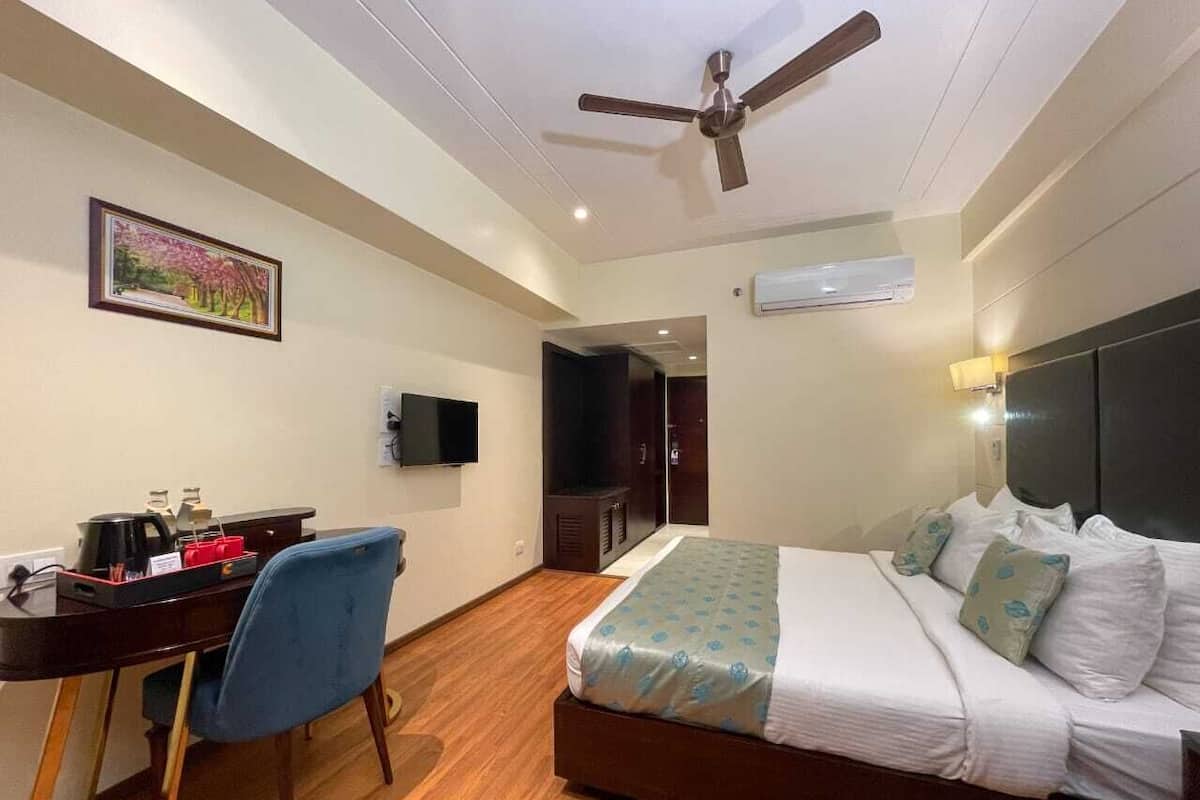 A hotel room with a bed, desk, chair, ceiling fan, TV, and an air conditioning unit.