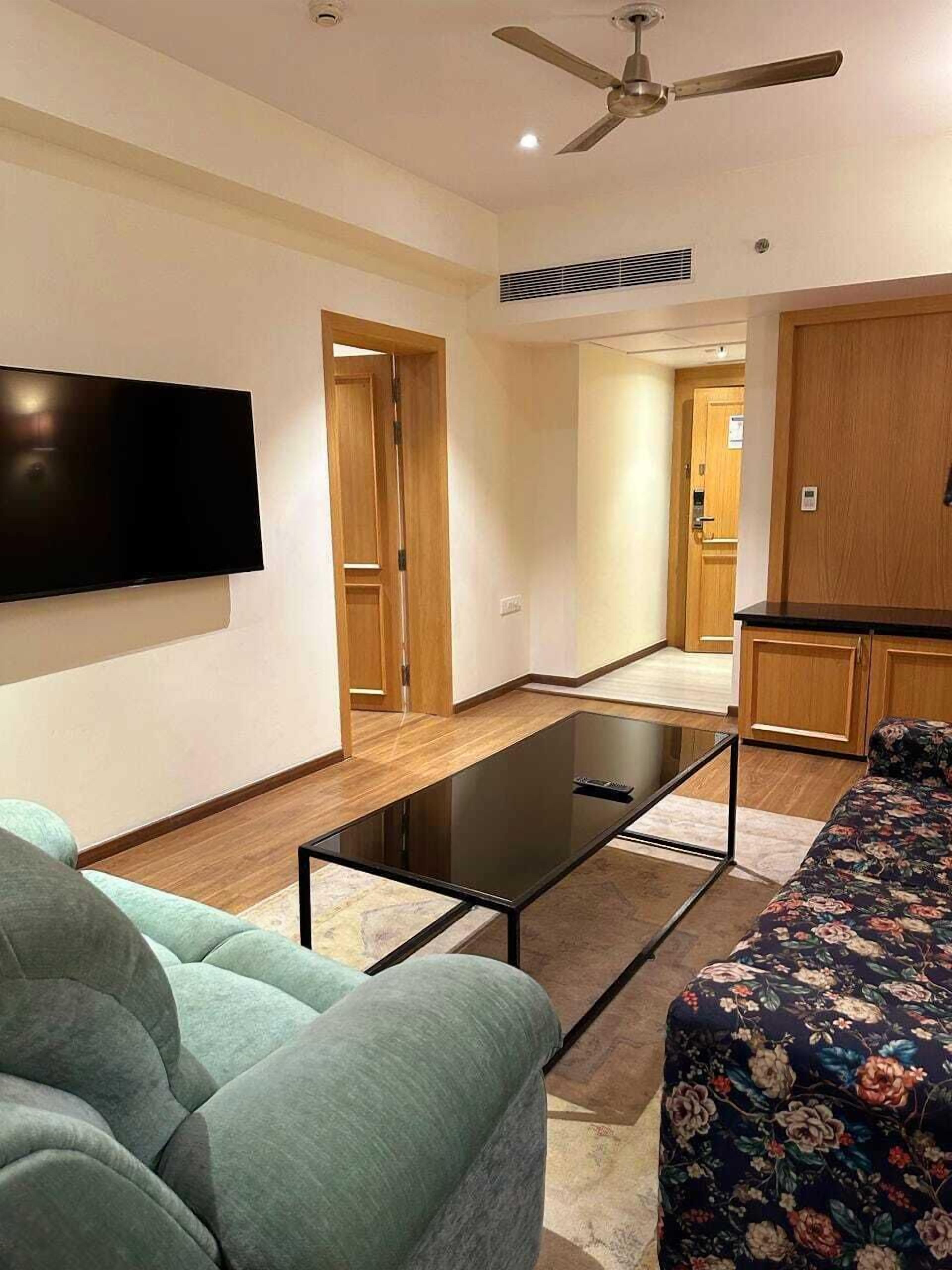 A modern living room with a flat-screen TV, a glass coffee table, a sofa, and a ceiling fan.