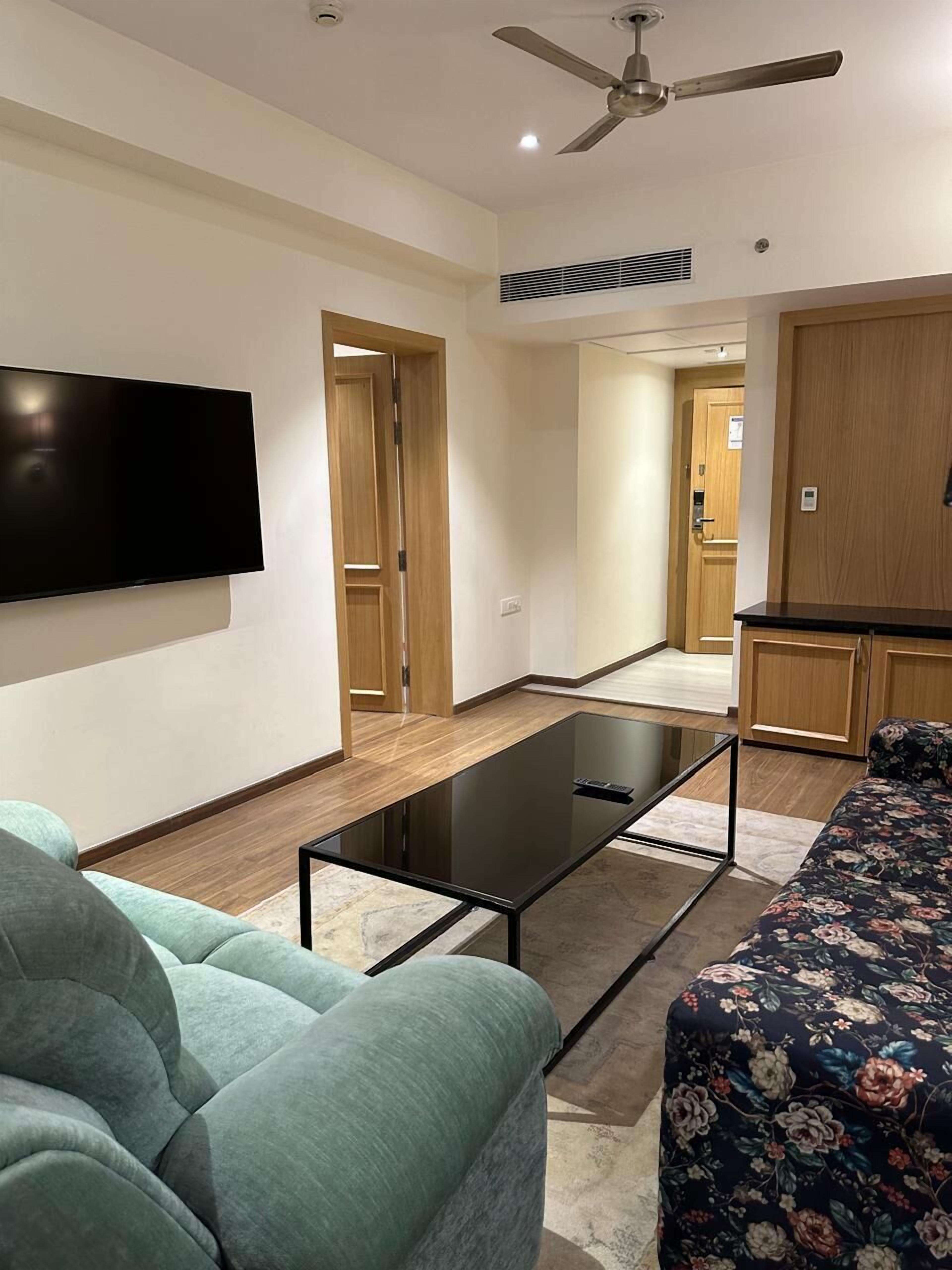 A modern living room with a flat-screen TV, a glass coffee table, a sofa, and a ceiling fan.