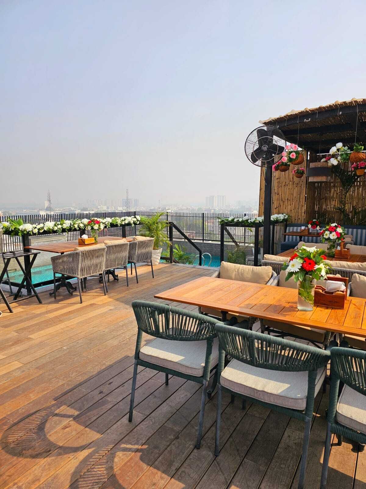 A rooftop bar with wooden tables and chairs, a pool, and a city view.
