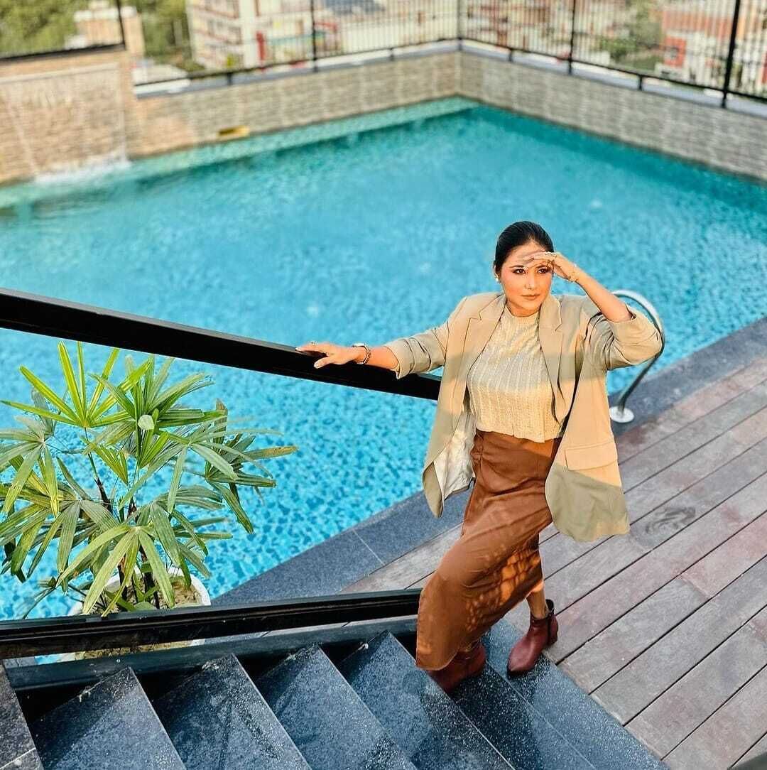 A person standing by a pool with a railing, surrounded by greenery and buildings in the background.