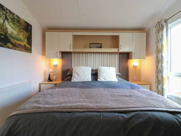 A bedroom with a bed, two bedside tables with lamps, a headboard with storage, and a window with curtains.