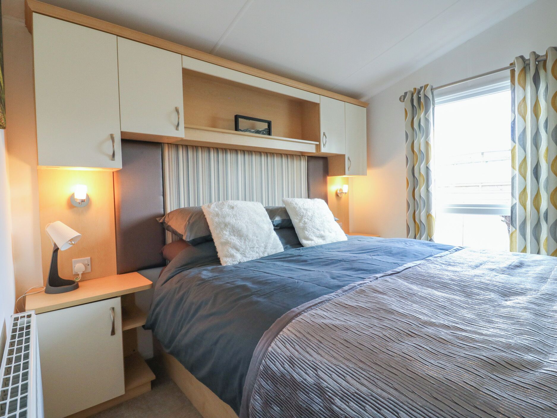 A bedroom with a bed, bedside lamps, a nightstand, a shelf with a picture, and a window with curtains.