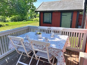En terrass med ett bord dukat för fyra personer, omgiven av vita stolar, och en liten röd byggnad i bakgrunden.