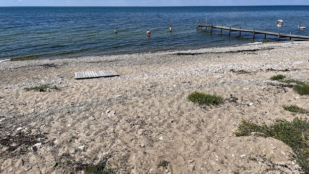 Kivikkoinen ranta, josta ulottuu tyynen veden ylle puinen laituri. Rannalla on pelastusrengas ja kasvillisuus on niukkaa.
