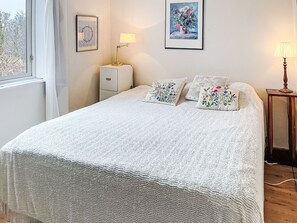 En snyggt bäddad säng med ett mönstrat, vitt täcke, två broderade kuddar, ett sängbord med en lampa, ett litet skåp och inramade konstverk på väggen.