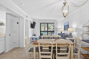 A dining area with a table and chairs, a TV mounted on the wall, and a view through a window. - Tennis Villa G6 - 2BR Beach Retreat Walk to Beach (Hilton Head Island)