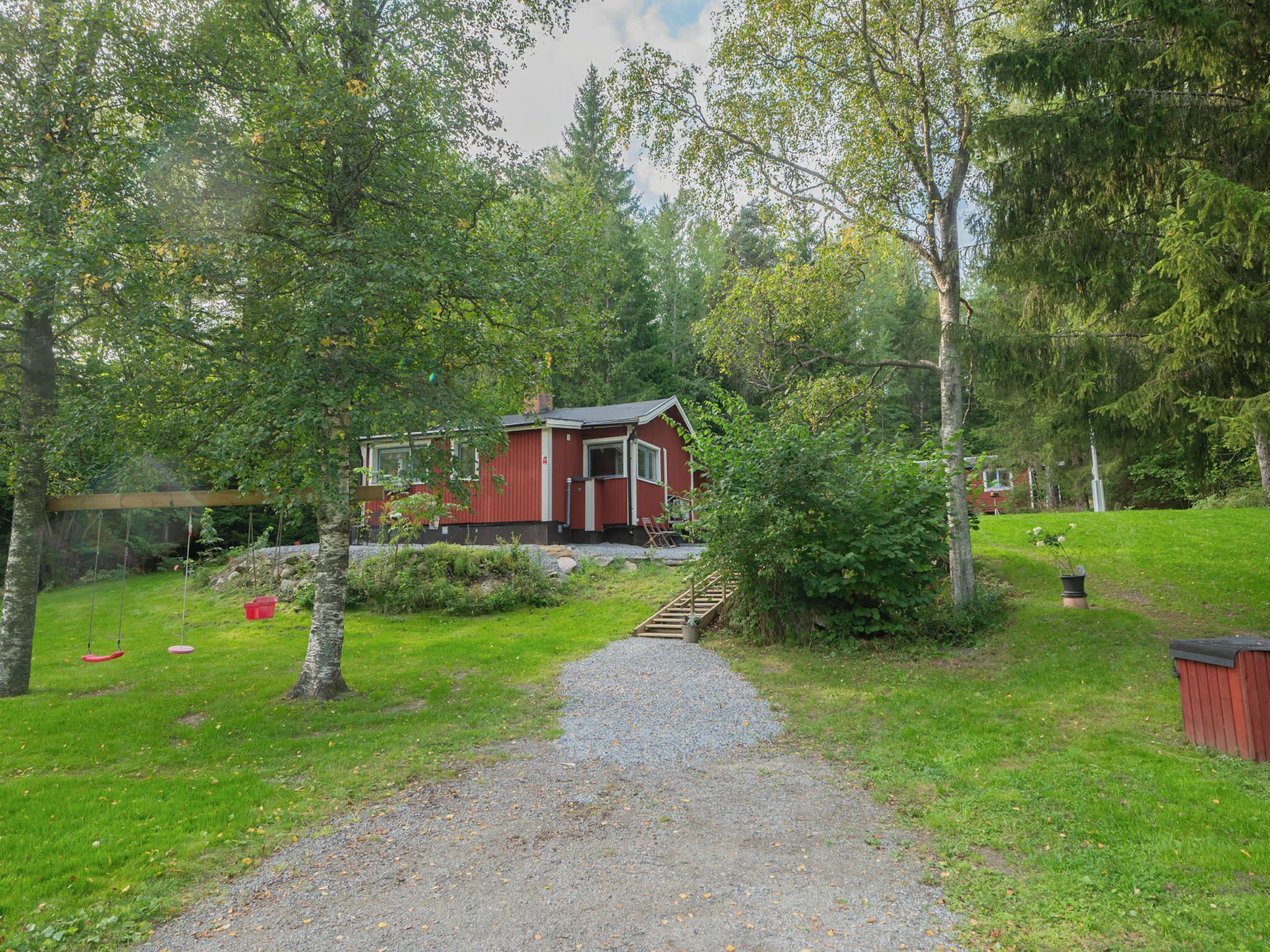 A red cabin with a gravel path leading to it, surrounded by greenery and trees.