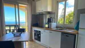 A modern kitchen with white cabinets, a wooden countertop, a built-in oven, a microwave, and a dishwasher. There is a dining table with chairs and a view of the ocean through large windows. - Peaceful lodging in Tampon, ocean view - 2 guests (Le Tampon)