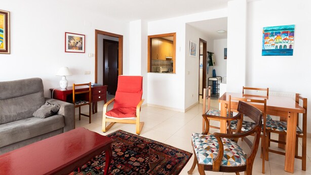 Ein Wohnzimmer mit einer Couch, einem roten Sessel, einem Esstisch mit Stühlen, einem gemusterten Teppich und einer durch einen offenen Durchgang einsehbaren Küche.