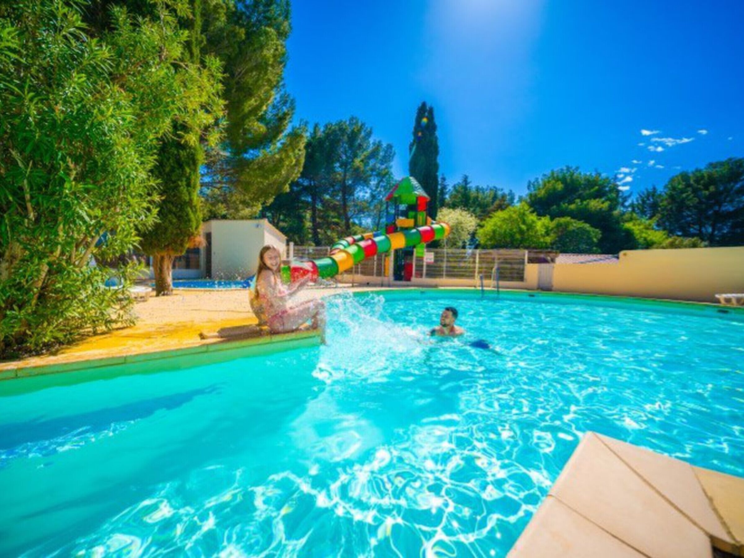 A swimming pool with a water slide, surrounded by trees and a clear sky.