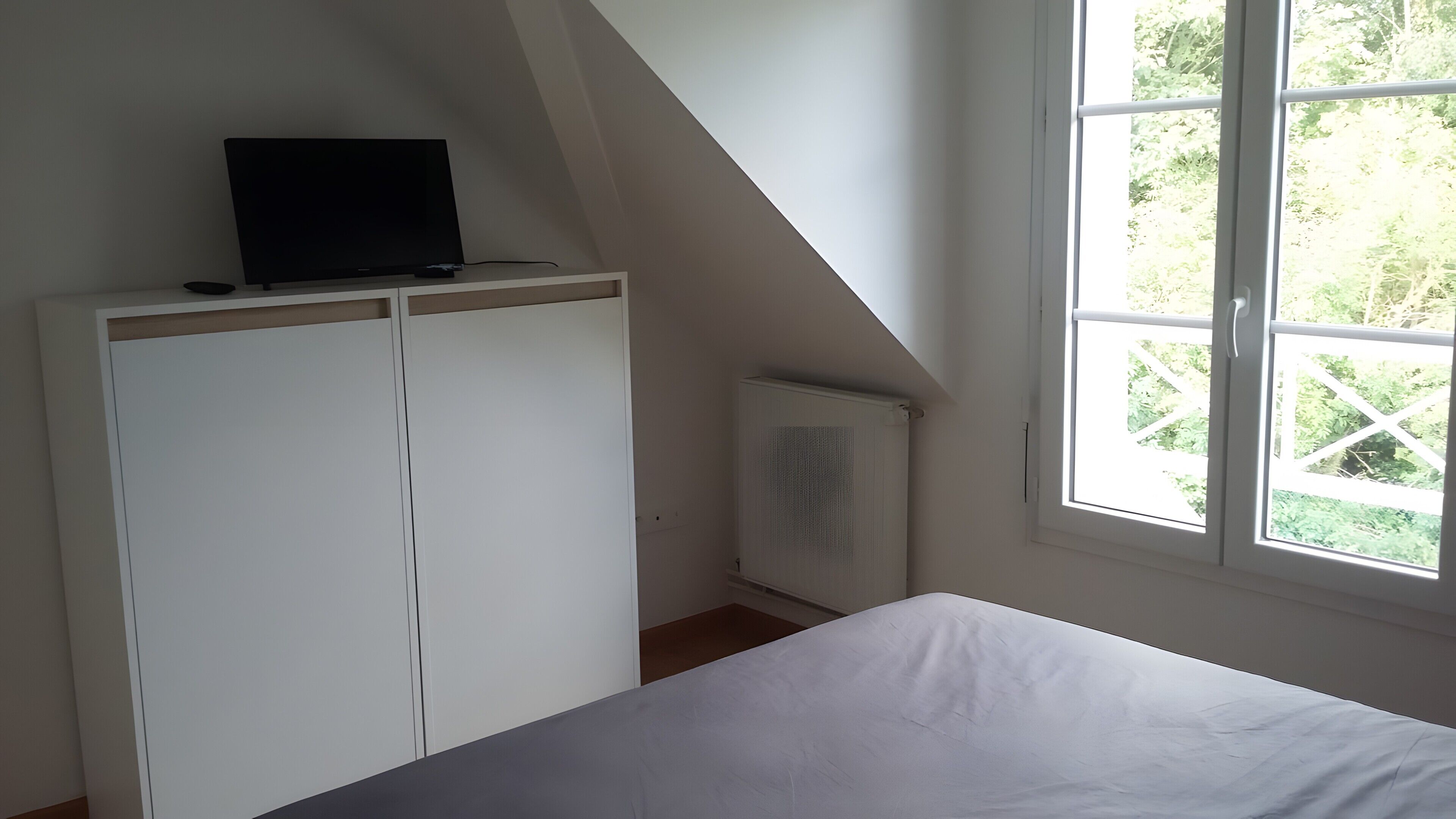Une chambre à coucher avec un lit, une armoire blanche, un radiateur et une fenêtre donnant sur un paysage verdoyant.