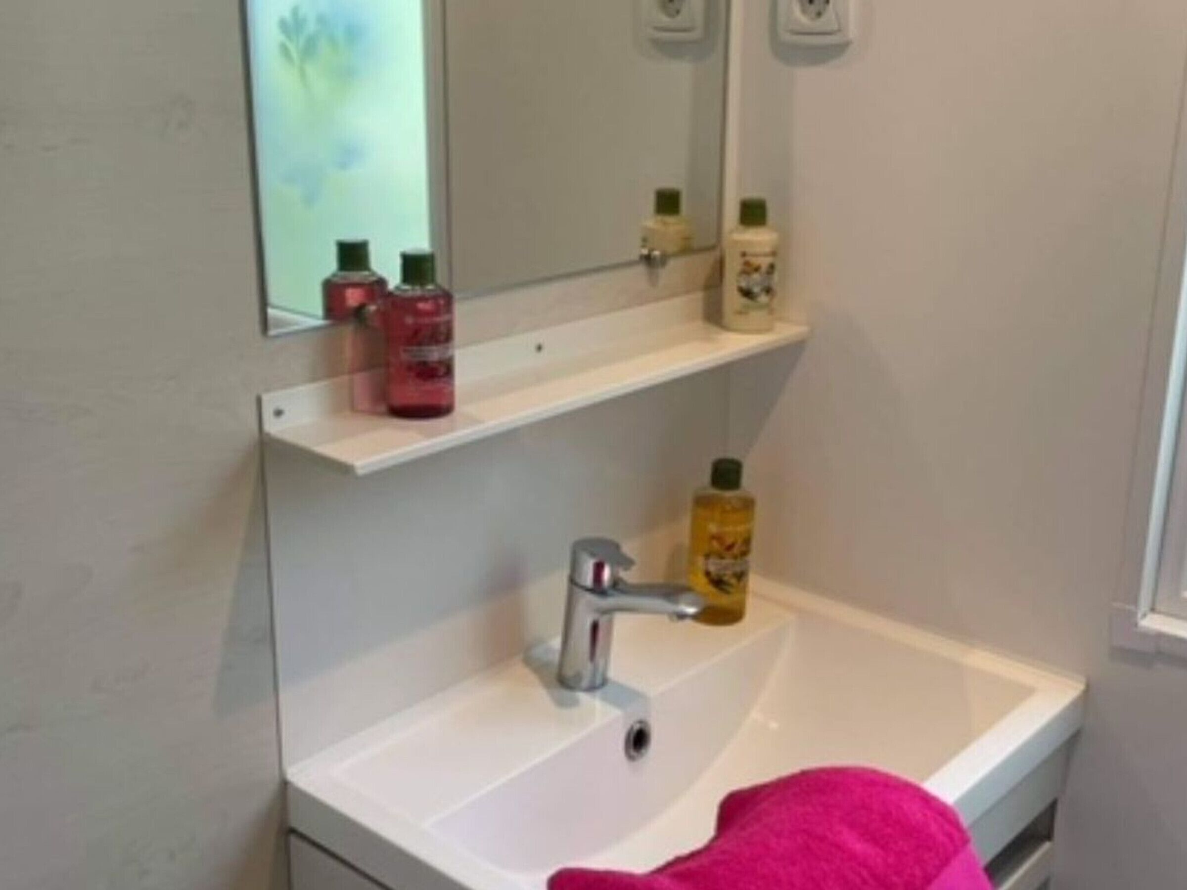 Un baño con lavamanos blanco, un espejo y dos estantes con artículos de aseo.