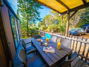 An outdoor deck with a table set for dining, surrounded by chairs, and a covered area with a yellow roof. - Air-conditioned mobile home - 4 guests (Balaruc-les-Bains)