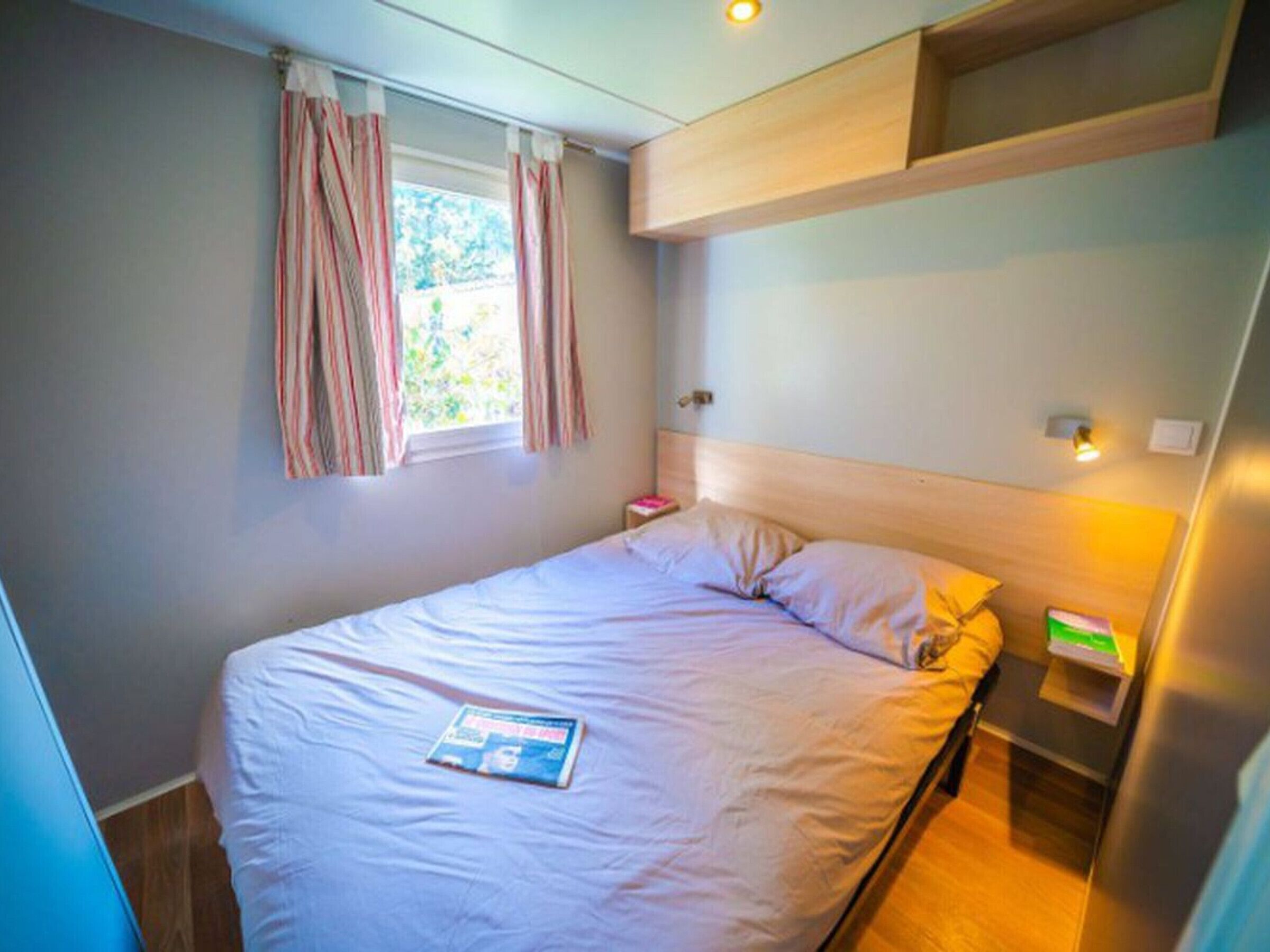 Et soveværelse med seng, gardiner ved vinduet, et sengebord med lampe og en hylde over sengen.