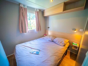 A bedroom with a bed, a window with curtains, a bedside table with a lamp, and a shelf above the bed. - Comfortable mobile home - 4 guests (Balaruc-les-Bains)