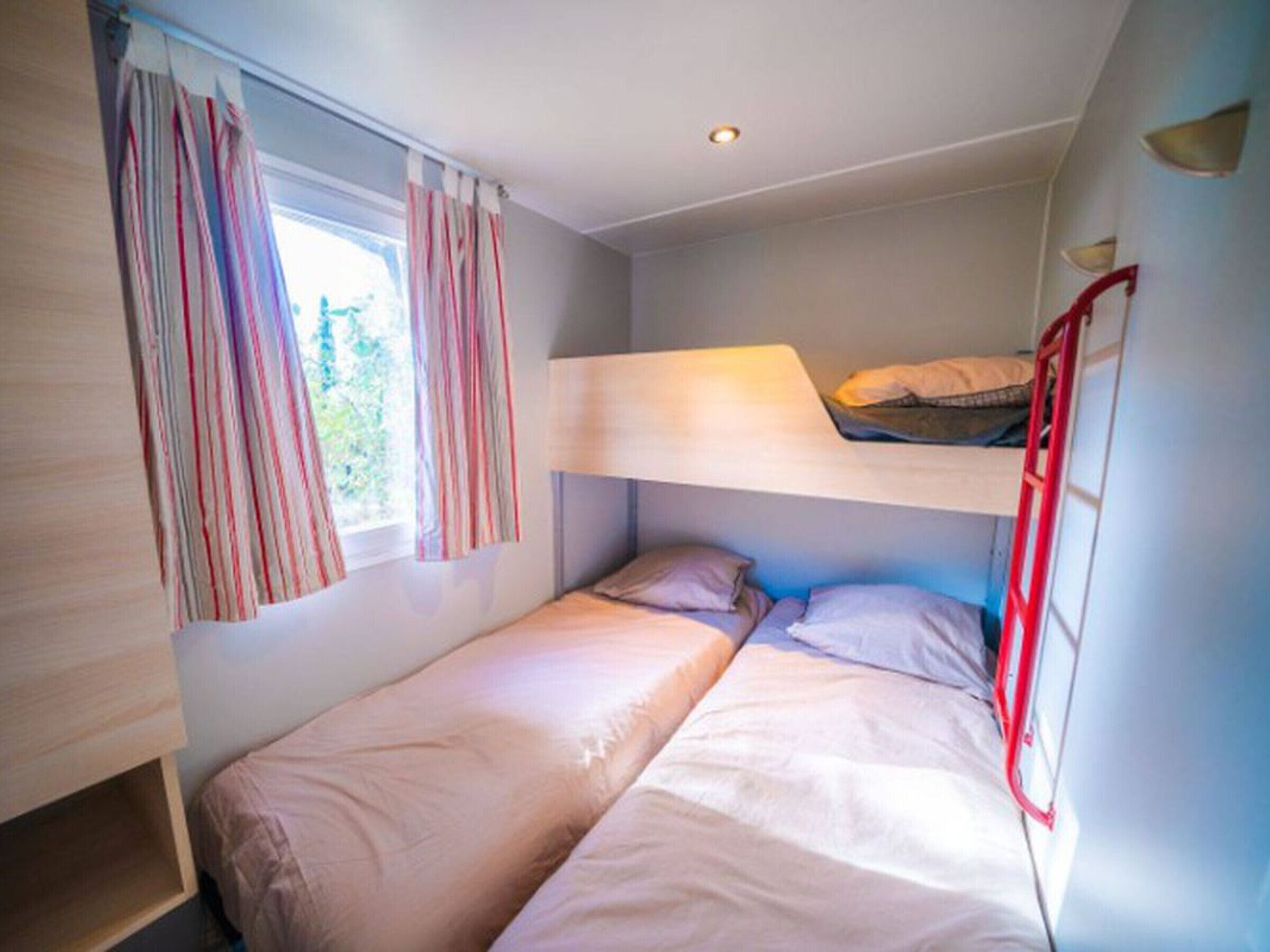 Ein kleines Zimmer mit Etagenbetten, einem Fenster mit Vorhängen und einer fest installierten Ablage.