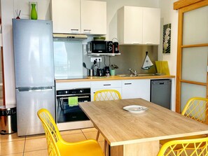A compact kitchen with a refrigerator, oven, microwave, and dishwasher. There is a dining table with chairs and a small shelf above the sink. - Lodging with pool, sea view - 4 guests (Le Tampon)