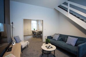 A modern living room with a sofa, coffee table, and TV. There is a staircase and a dining area in the background. - Vacant Nests | The Freda | Modern 3BR Home | (Sunderland)