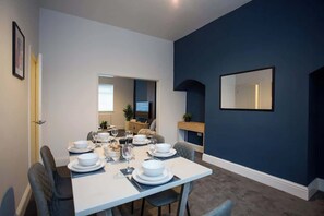 A dining area with a table set for four, a television in the background, and a dark blue accent wall. - Vacant Nests | The Freda | Modern 3BR Home | (Sunderland)
