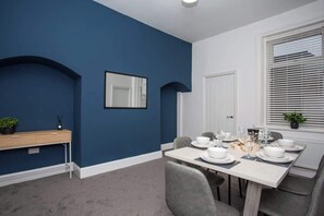 A dining area with a table set for four, a mirror, a white door, and a window with blinds. - Vacant Nests | The Freda | Modern 3BR Home | (Sunderland)
