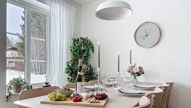 Un comedor con una mesa preparada para cuatro, una planta en maceta, velas y un reloj en la pared.