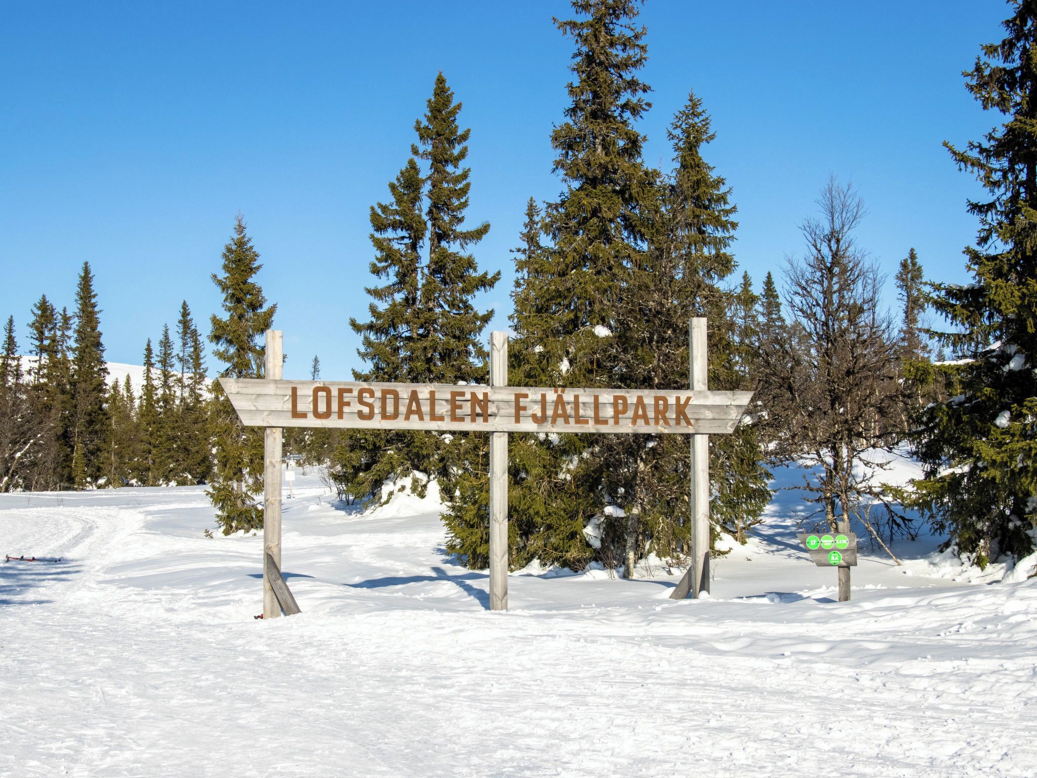Ein hölzernes Schild mit der Aufschrift 'LOFSDALEN FjALLpark', umgeben von Schnee und Bäumen.