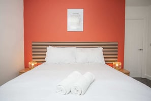 Una cama bien tendida con sábanas blancas y dos toallas enrolladas, un cuadro en la pared y lámparas de noche sobre mesitas de madera.