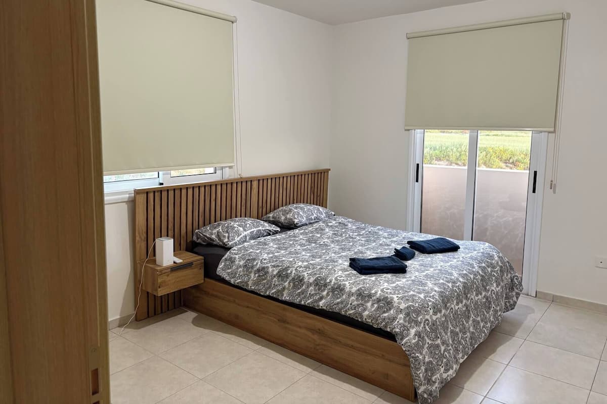 Une chambre à coucher comprenant un lit, une table de chevet, une fenêtre avec un store et une porte-fenêtre coulissante donnant sur un balcon.