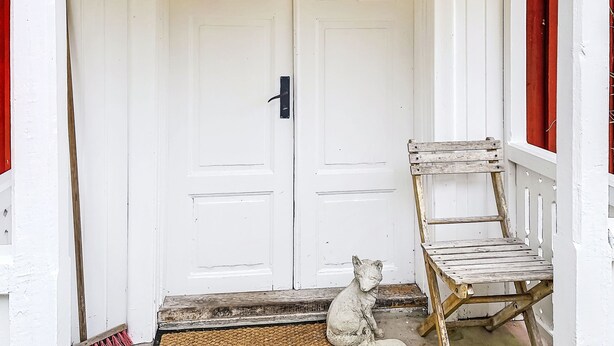Una puerta blanca con un ventanuco, un banco de madera y una estatua de piedra de un gato.